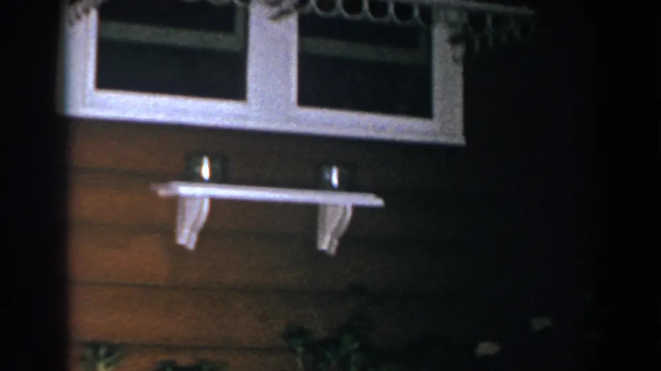 1960 home-movie view of a white-framed window, small exterior shelf, and brown siding above garden plants.