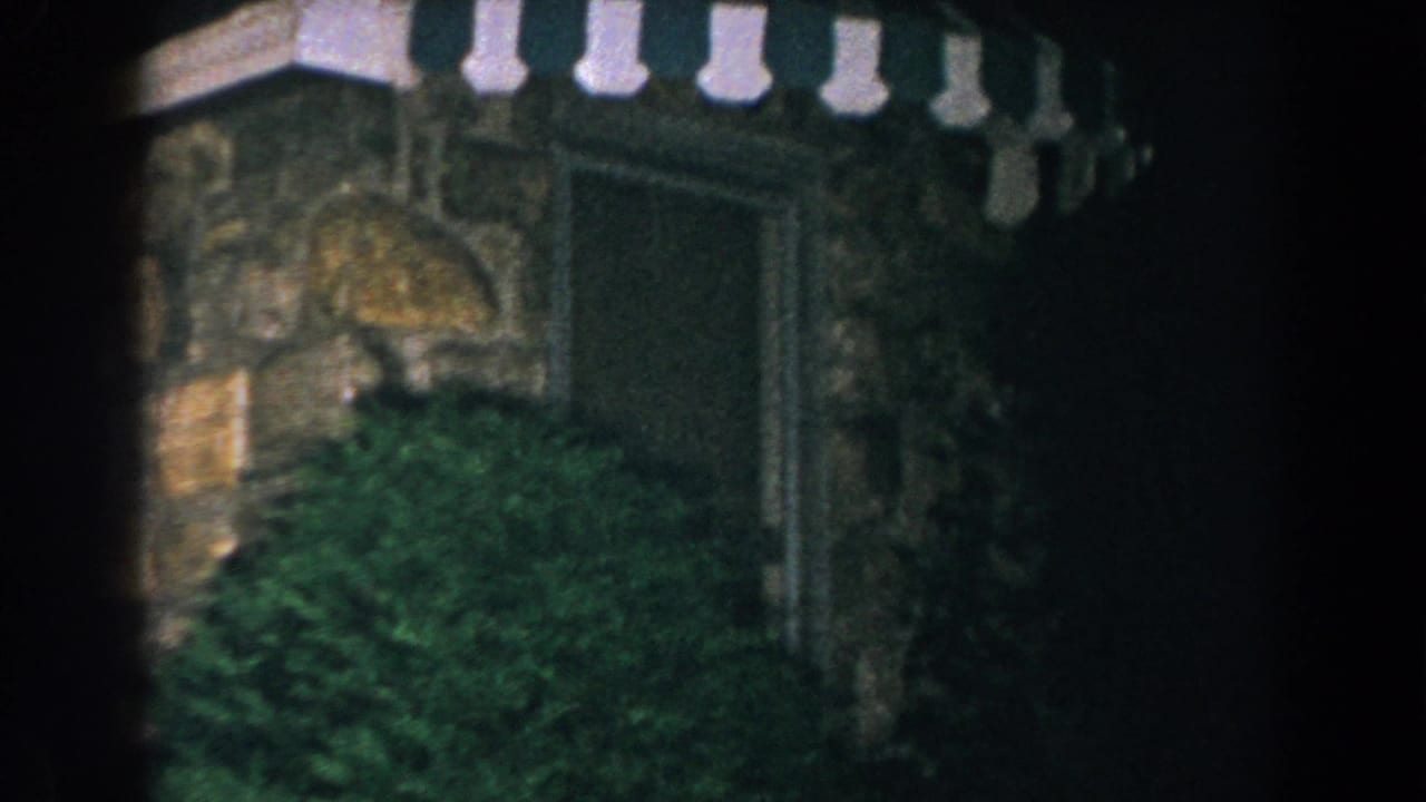 1960 Brighton, Michigan 8mm frame showing a dark stone exterior, striped awning, recessed doorway, and dense greenery.