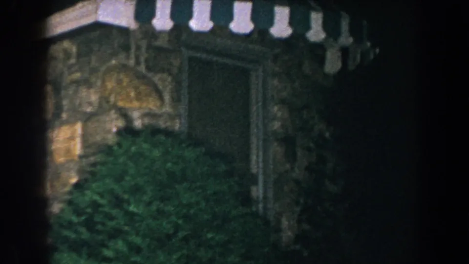 1960 Brighton, Michigan 8mm frame showing a dark stone exterior, striped awning, recessed doorway, and dense greenery.