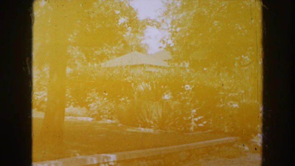 Yellowed 8mm outdoor frame from the Brighton, Michigan clip, showing trees, lawn, plantings, a pale tent-like or roof shape, dust, and gate shadows.