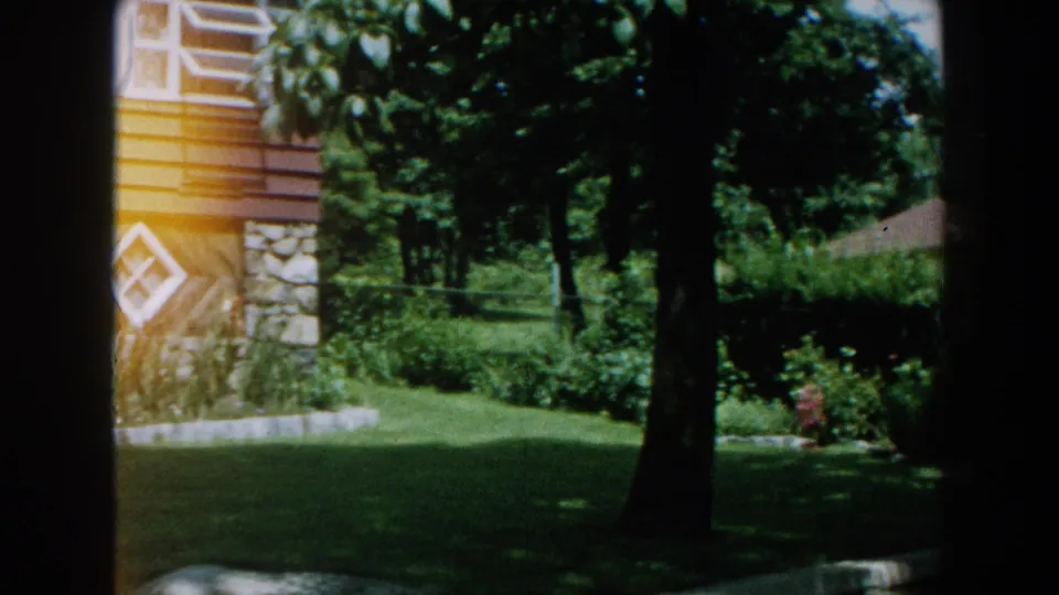 1960 8mm frame in Brighton, Michigan showing part of a suburban house with stone facade details, diamond window, trees, lawn, and garden plantings.