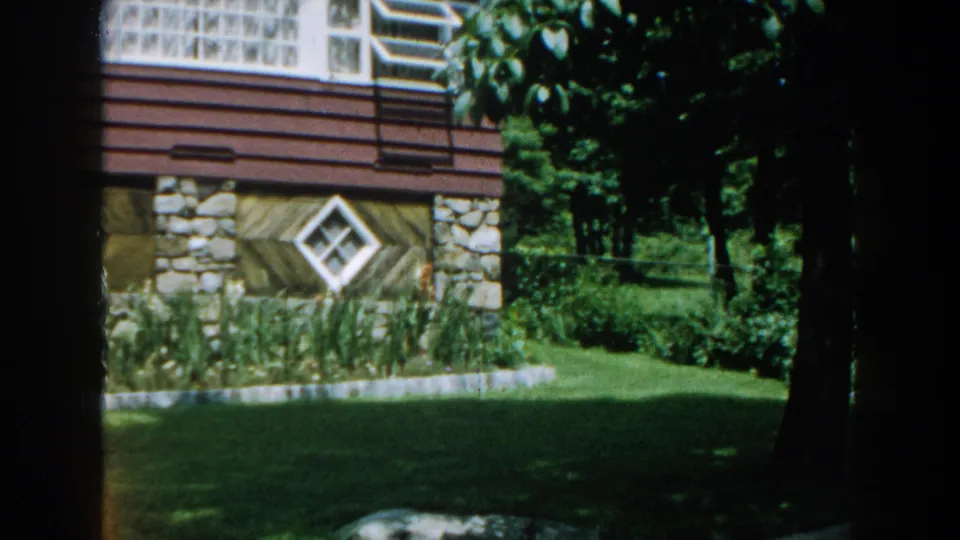 Late frame from the 1960 Brighton, Michigan clip showing the house exterior, stone base, diamond window, upper windows, trees, lawn, and garden beds.