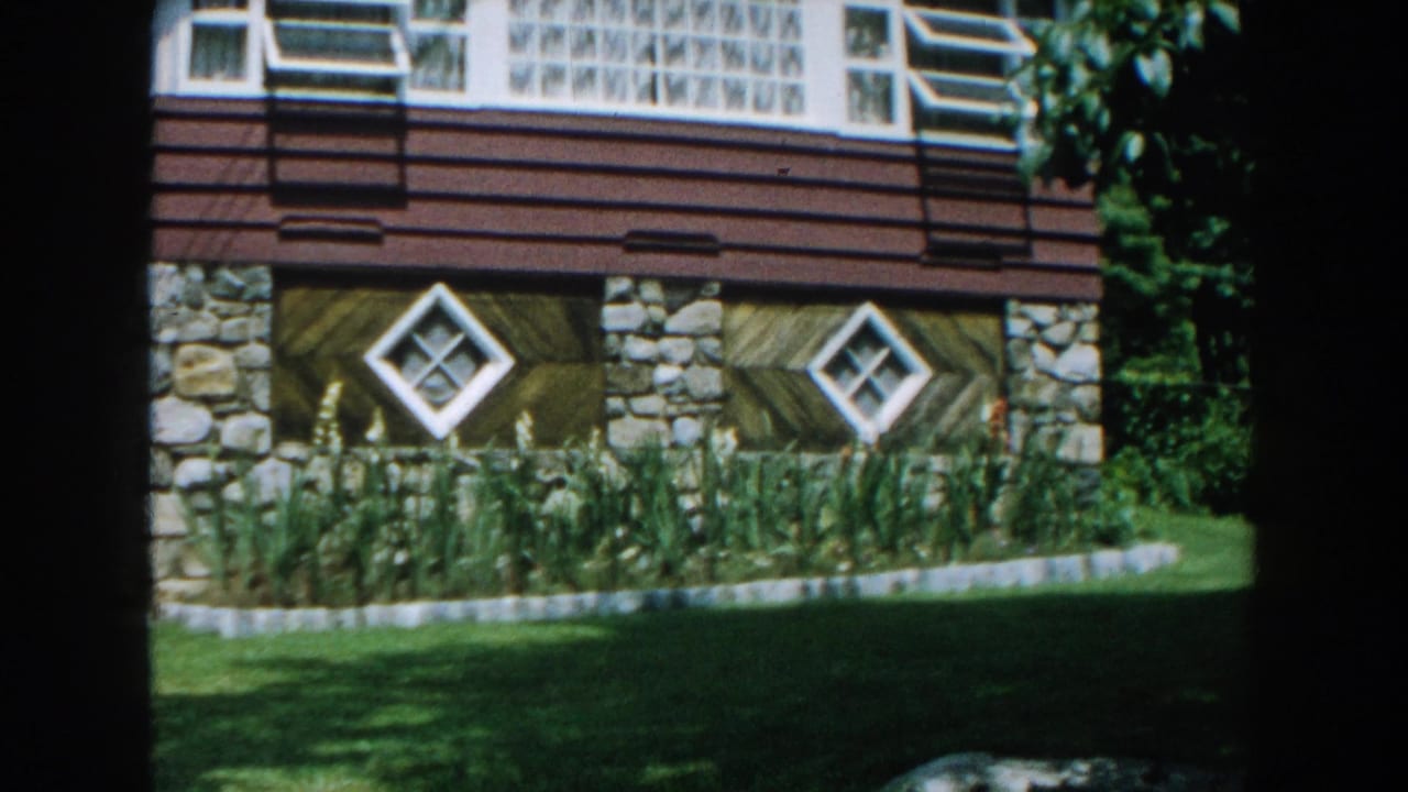 House facade with brown siding, stone supports, diamond windows, lawn, trees, and flower bed.