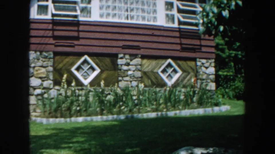 Brighton, Michigan 1960 frame with the house centered behind lawn and tall plantings, showing stone supports and diamond-shaped windows.