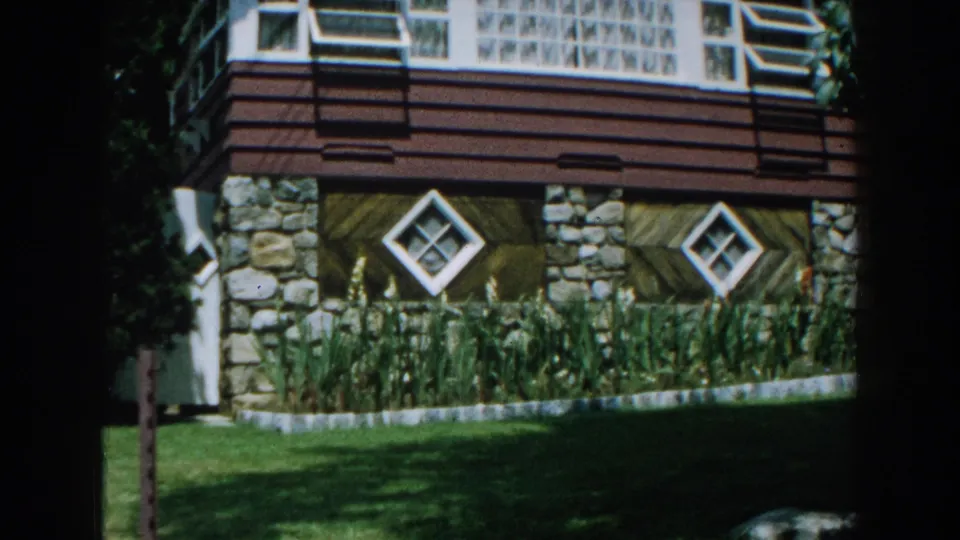 Closer 1960 home-movie view of the Brighton house wall, with stone columns, diamond windows, tall plants, and a shaded lawn edge.