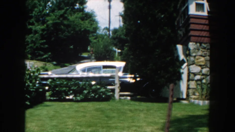 A white-roof sedan passes a manicured Brighton, Michigan yard, with shrubs, a white post, and house exterior visible.