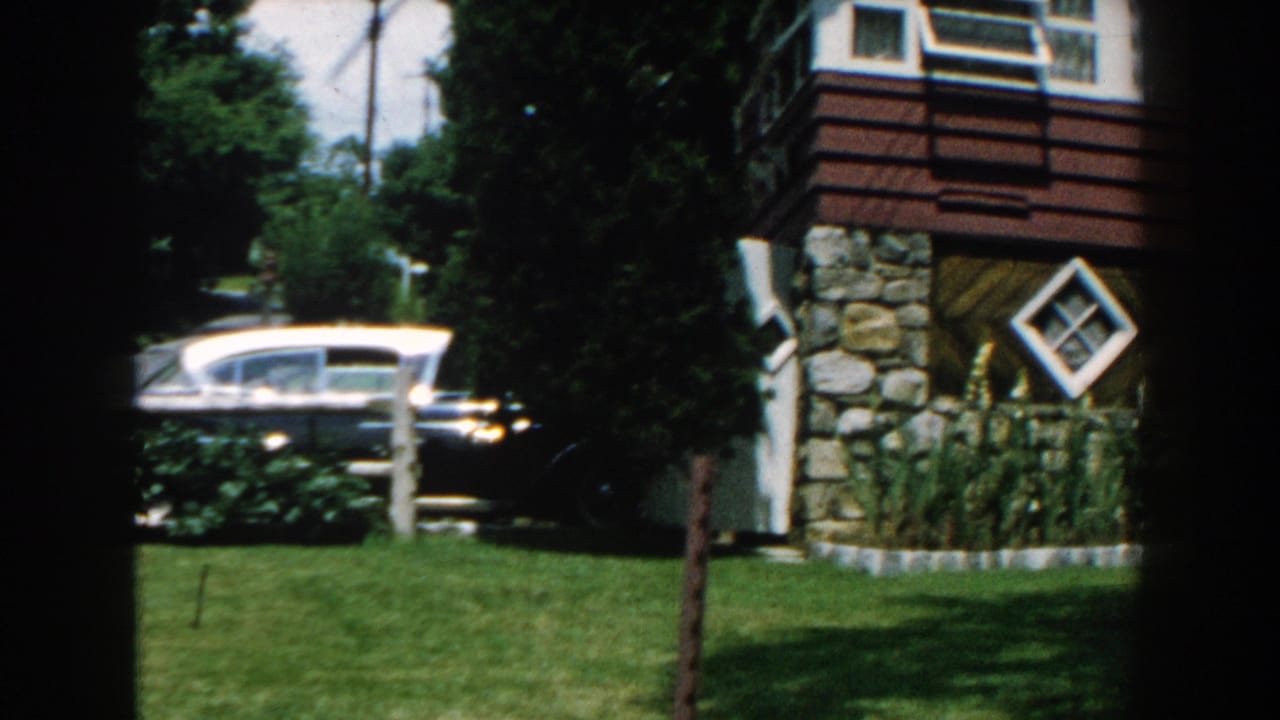 Suburban house exterior with lawn, trees, and a parked period car beside the building.