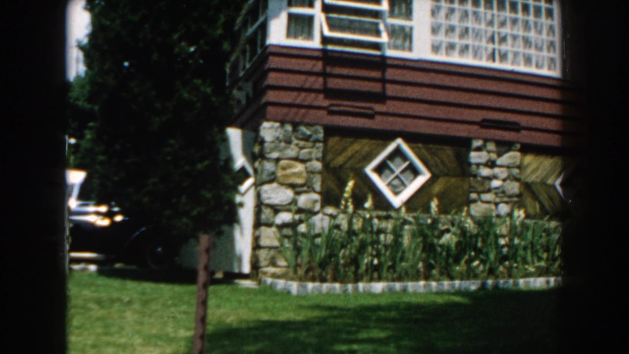 House corner with stone base, brown siding, diamond window, lawn, and a parked car at left.