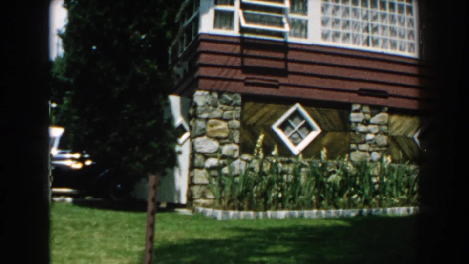1960 Brighton, Michigan frame of a suburban house corner with stonework, diamond window, lawn, trees, and a partly seen car.