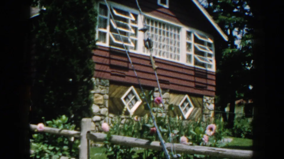 1960 Brighton, Michigan home-movie frame showing a house facade with diamond windows, flowers, fence, lawn, and tree shade.