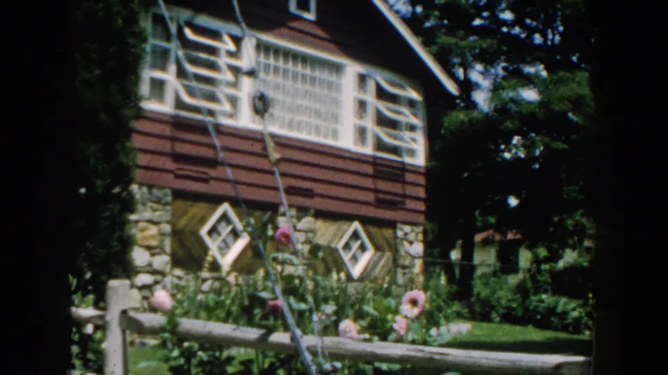 1960 Brighton, Michigan frame of a sunlit suburban residence with stone base, brown siding, diamond windows, flower bed, and fence.