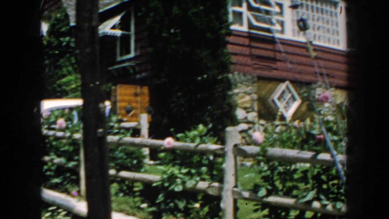 House corner and garden partly obscured by dark vertical film edges and trees.