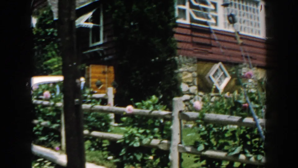 1960 Brighton, Michigan view of the house corner, greenery, fence, and driveway area partly framed by dark film borders.