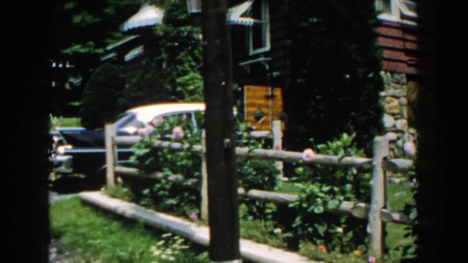 1960 Brighton, Michigan frame with a parked period car beside the rustic house, garden bed, split-rail fence, and trees.