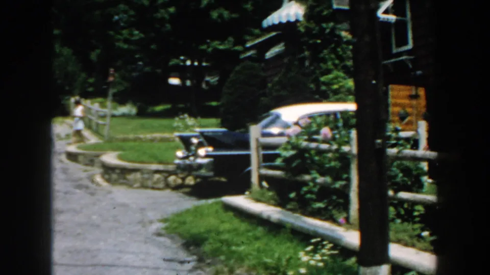 1960 Brighton, Michigan view down a curved driveway past a split-rail fence, pink flowers, house exterior, and parked car.