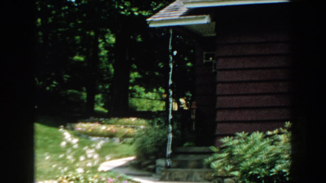 1960 Brighton, Michigan frame with a brown-sided house exterior, bright lawn, flower beds, shrubs, and dense tree shade.