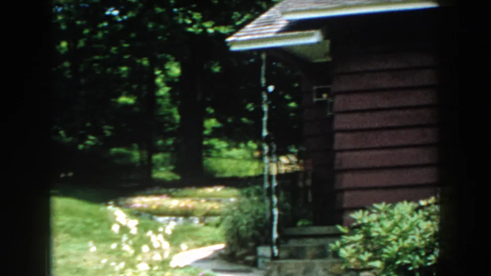 1960 Brighton, Michigan home-movie frame showing the house wall, shaded porch area, lawn, blooming flowers, shrubs, and trees.
