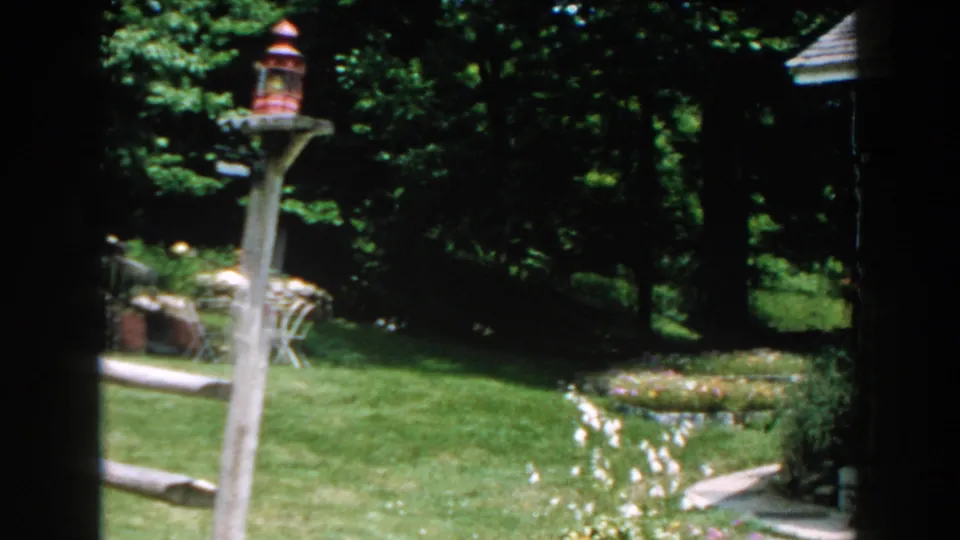 1960 Brighton, Michigan frame with a red bird feeder on a wooden post, lawn, flower beds, trees, and the edge of a brown house.