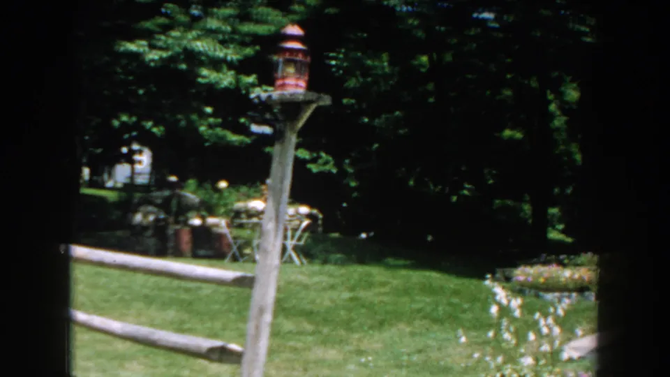 1960 Brighton, Michigan backyard view with a rustic fence, decorative lantern post, green lawn, trees, and distant patio furniture.
