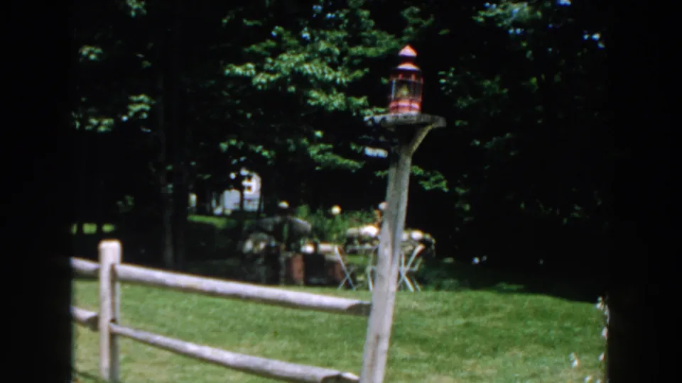 1960 Brighton, Michigan home-movie frame showing a backyard fence, lantern post, shaded lawn, trees, and patio furniture.