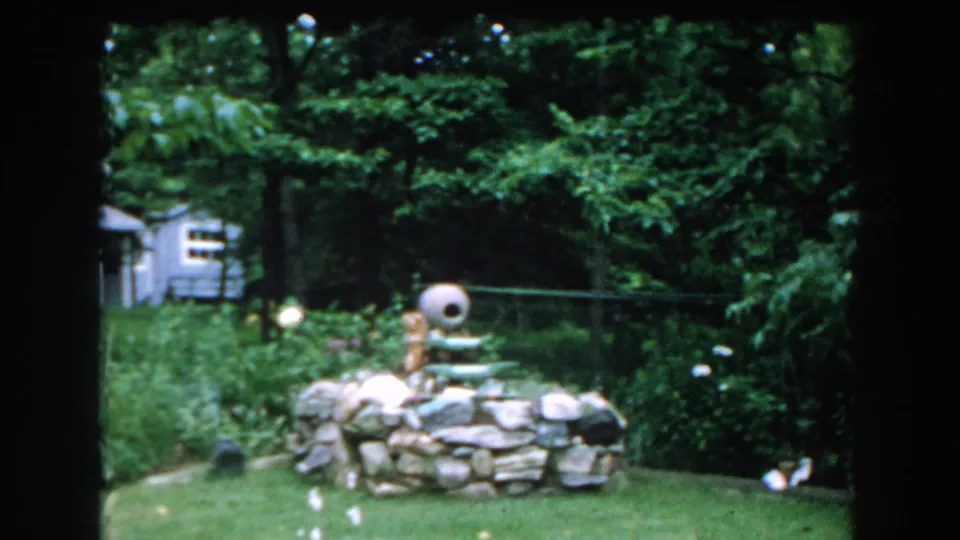 1960 Brighton, Michigan backyard view with a stone fountain, lawn, garden plantings, trees, and a small shed in the background.