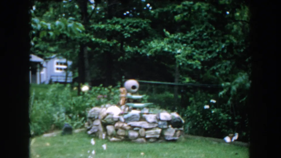 1960 Brighton, Michigan frame showing a backyard stone fountain, surrounding lawn, shrubs, trees, flowers, and a small shed.