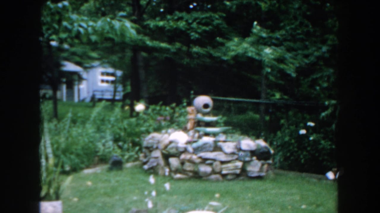 Backyard garden with stone water feature, lawn, trees, and small blue structure in the background.