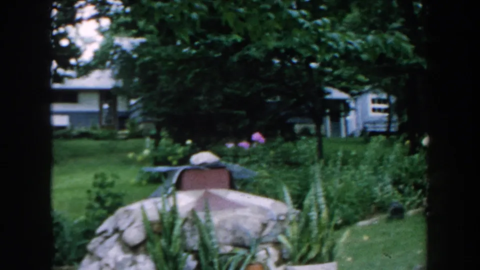 1960 Brighton, Michigan garden frame showing a lawn, covered object, leafy trees, tall plantings, and a small blue structure behind the yard.
