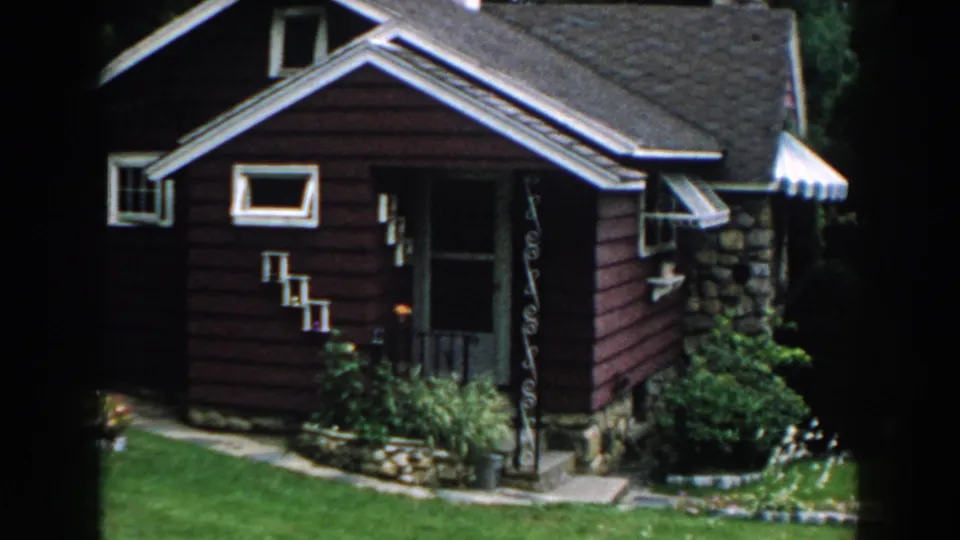 1960 Brighton, Michigan home-movie frame showing a red bungalow with white trim, stone base, striped awning, shrubs, and lawn.