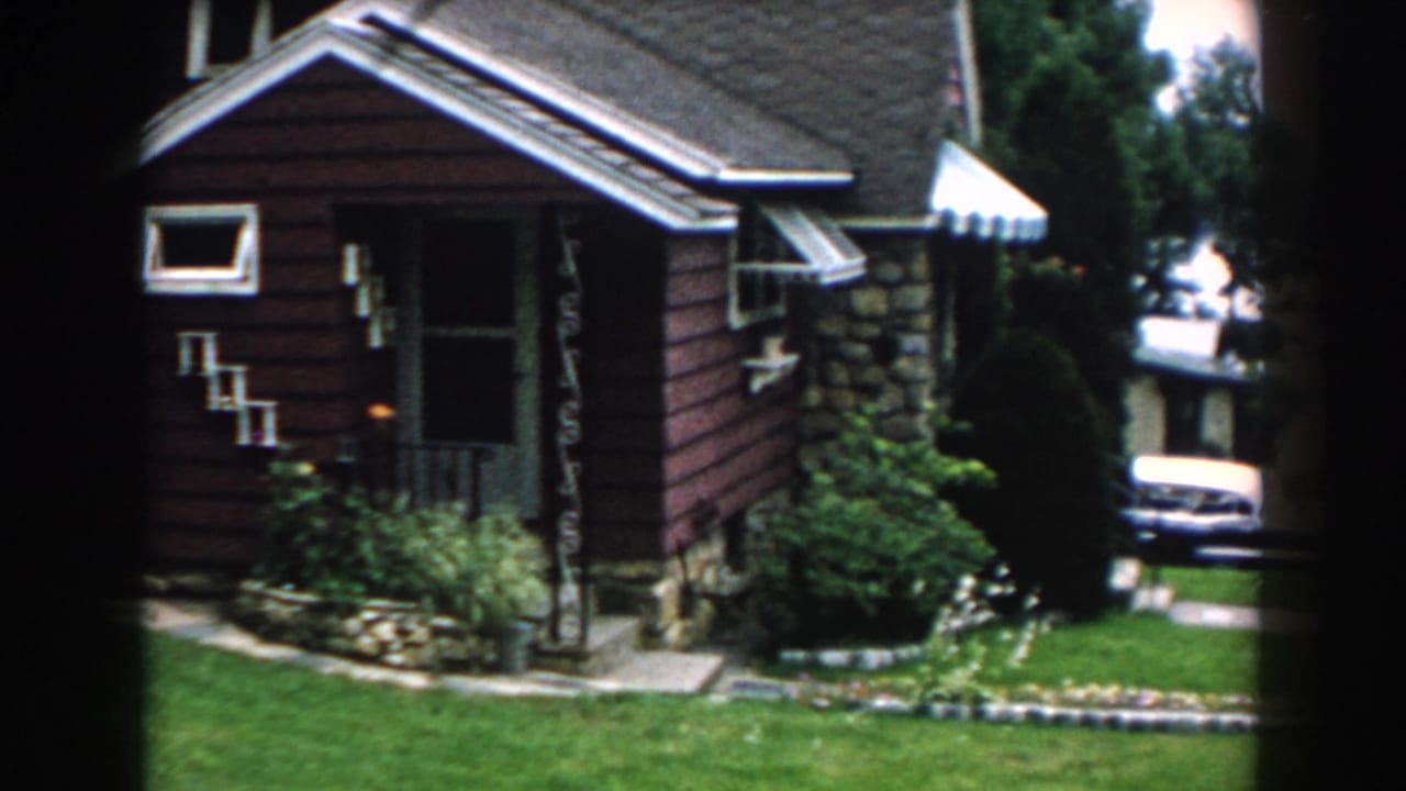 Front of the bungalow with awning, shrubs, lawn, and a car partly visible at right.