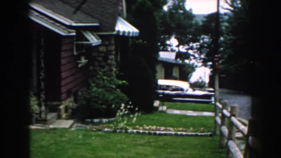 1960 Brighton, Michigan home-movie frame showing the side of the red bungalow, garden edge, lawn, trees, and a parked car beyond.