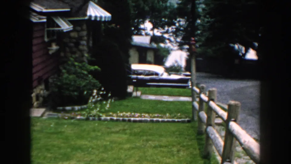 1960 Brighton, Michigan frame with the house edge, green lawn, split-rail fence, driveway, parked car, shrubs, and trees.