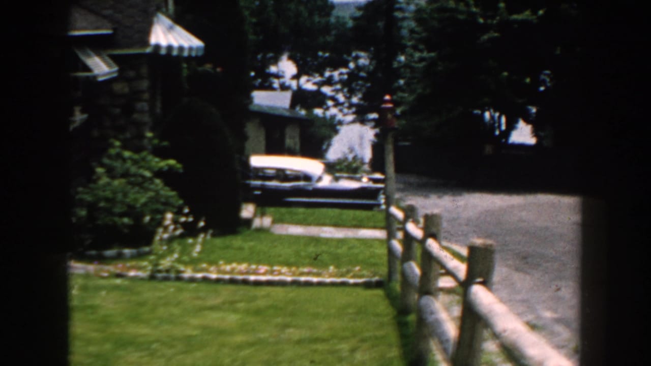 1960 Brighton, Michigan home-movie frame showing a green lawn, driveway, stone house, trees, fence, and a classic two-tone car in the background.
