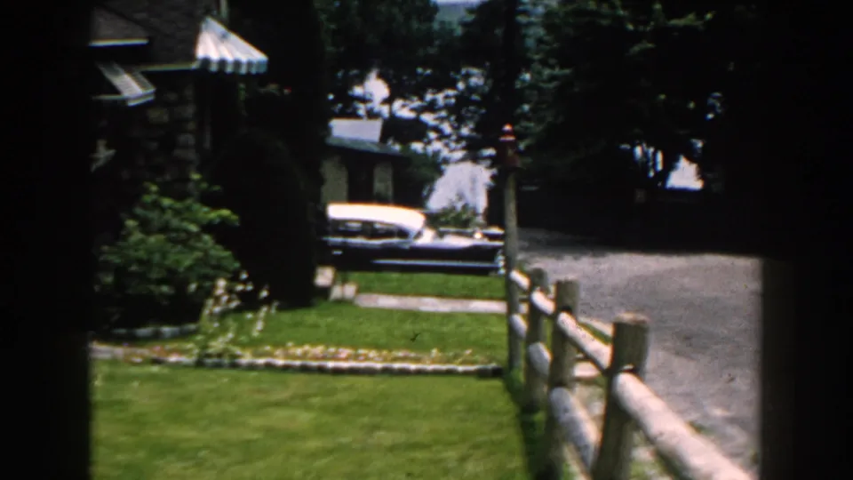 1960 Brighton, Michigan home-movie frame showing a green lawn, driveway, stone house, trees, fence, and a classic two-tone car in the background.