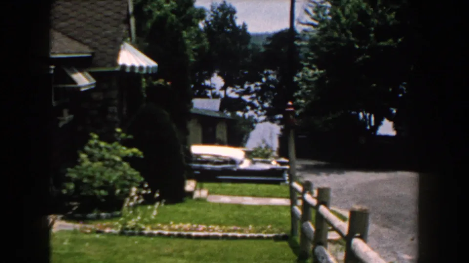 1960 Brighton, Michigan exterior view with a two-tone classic car parked near a driveway, bordered by lawn, fence, trees, and a stone house.