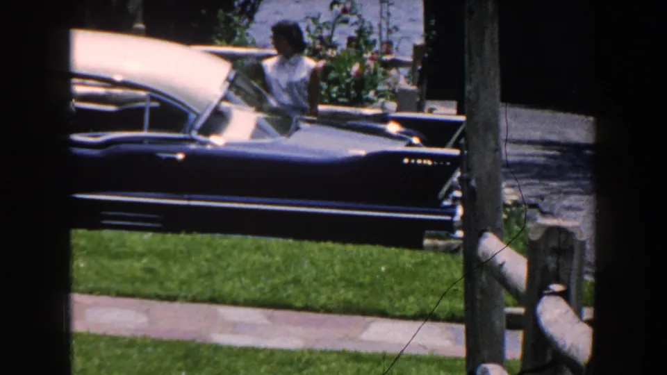 1960 Brighton, Michigan frame showing a woman in a light sleeveless top beside a two-tone classic car near a lawn, fence, and driveway.