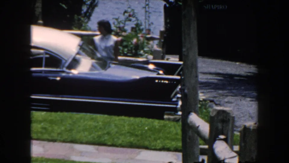 1960 Brighton, Michigan home-movie frame with a woman beside a parked two-tone car, seen across a lawn with fence posts and garden planting.