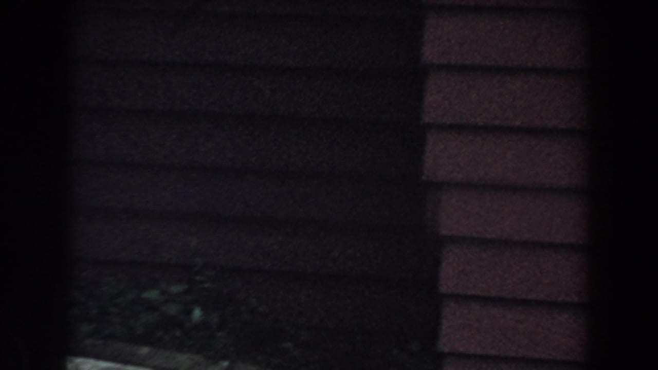 1960 Brighton, Michigan 8mm frame showing a shaded house exterior with horizontal siding and dark garden foreground.