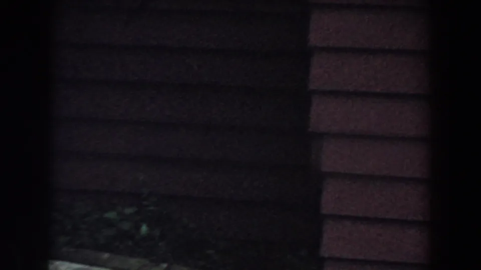 1960 Brighton, Michigan 8mm frame showing a shaded house exterior with horizontal siding and dark garden foreground.
