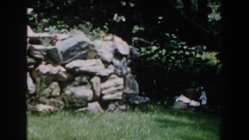 1960 Brighton, Michigan home-movie frame showing a sunlit garden, rough stone wall, lawn, and shaded foliage.