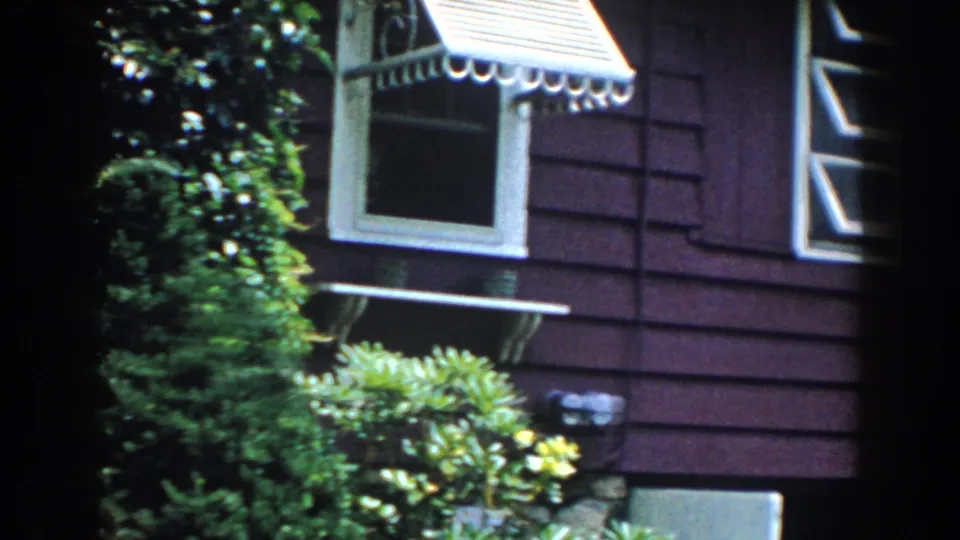 1960 Brighton, Michigan frame showing red siding, white-trimmed windows, a striped awning, shrubs, ivy, and house foundation details.