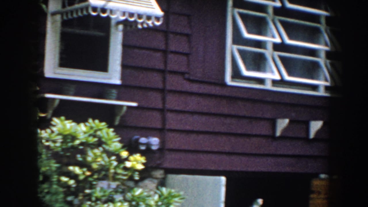 1960 Brighton, Michigan 8mm frame showing a maroon-sided suburban house exterior with white trim, a striped awning, shrubs, and foundation stonework.
