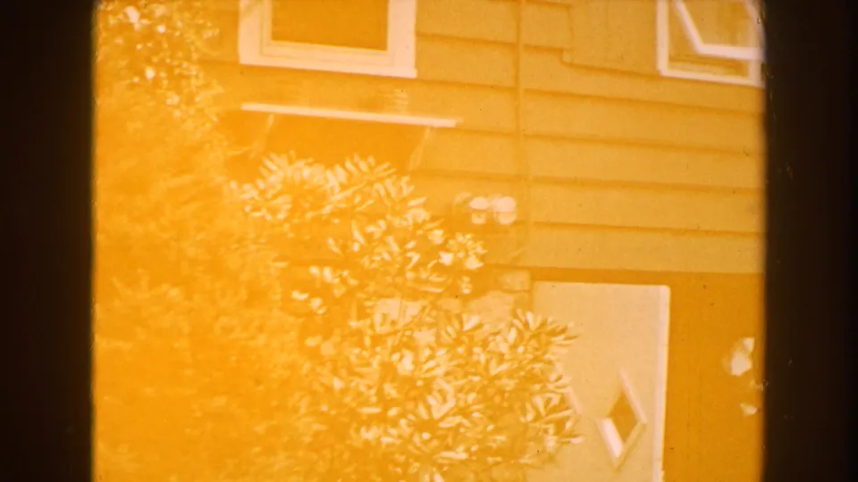 Light-leaked 1960 Brighton, Michigan 8mm frame where the house siding, window trim, shrubs, and foundation remain faintly visible.