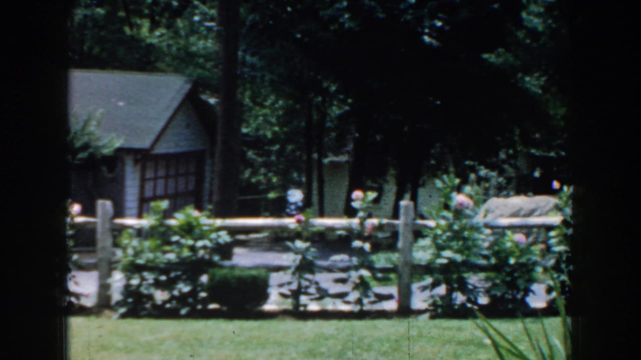 Small house behind a white fence, stone edging, shrubs, and trees.