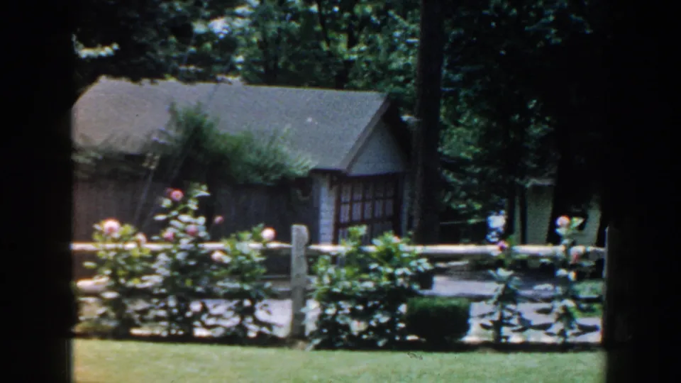 1960 home-movie frame of a Brighton, Michigan garden with a small house, blooming shrubs, fence posts, and trimmed grass.