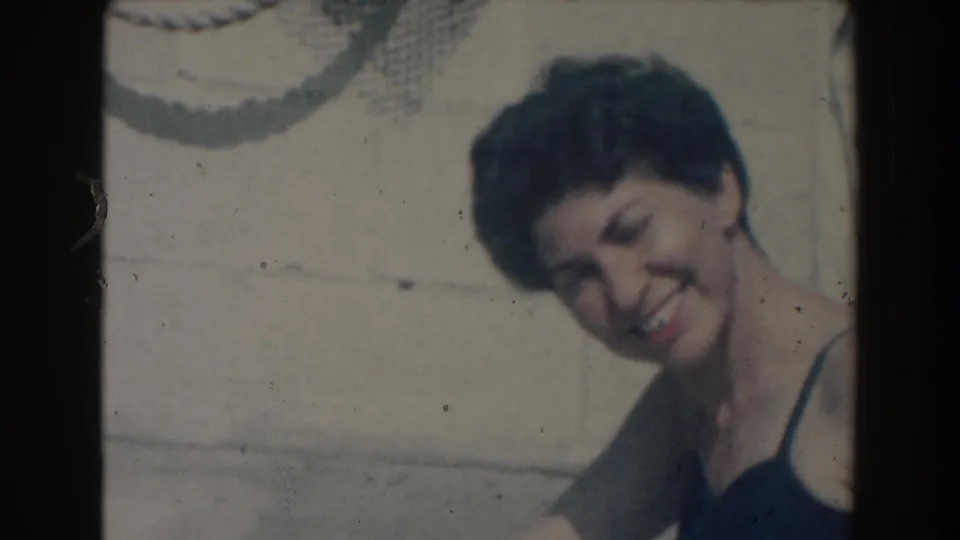 In 1961 North Hollywood home-movie footage, a woman in a dark swimsuit smiles toward the camera near a swimming pool.