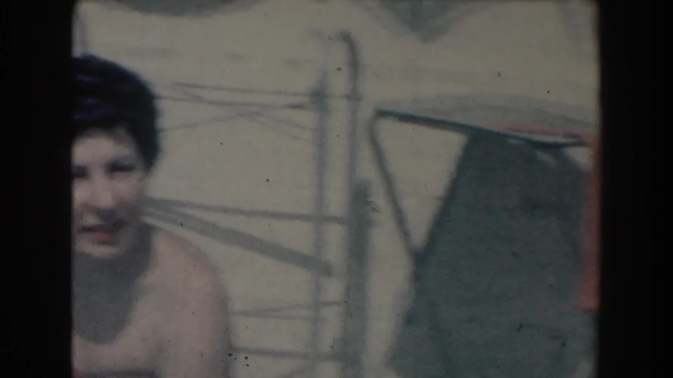 A woman appears low in the frame near a swimming pool, partly obscured by chair legs and the soft home-movie exposure.