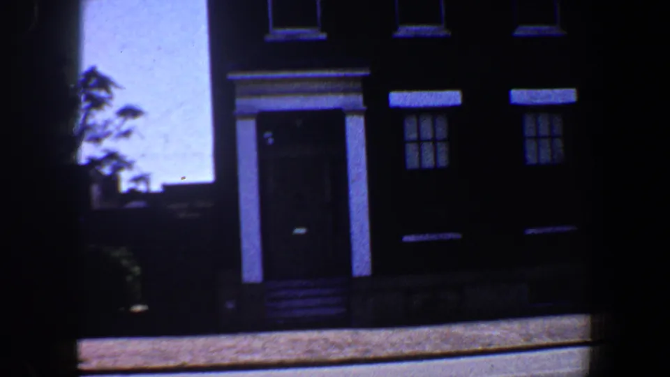 1964 Saint Louis 8mm frame showing a dark three-story residential facade with white trim, a central doorway, front steps, and bright sky.