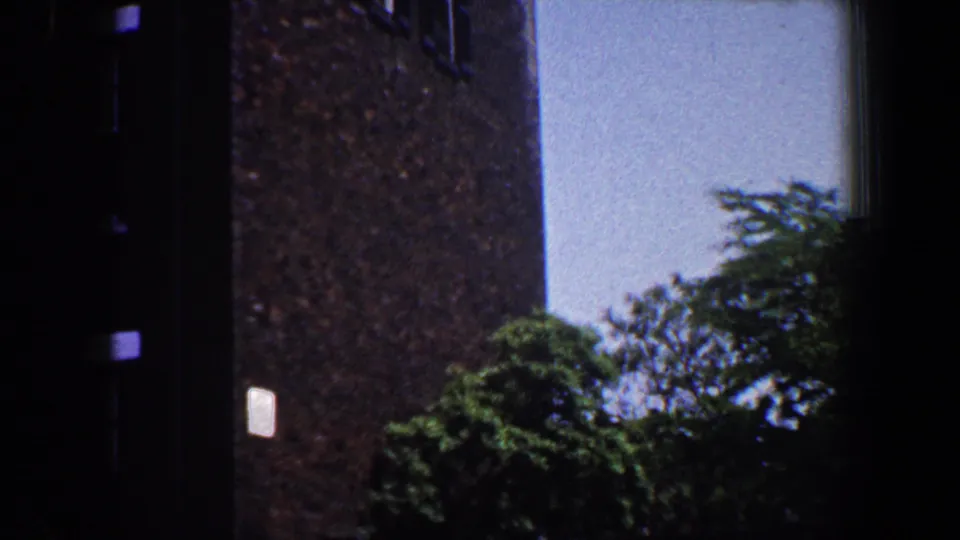 1964 Saint Louis exterior frame with a tall brown brick wall, pale side wall, trees, and a small light-colored sign or plaque.