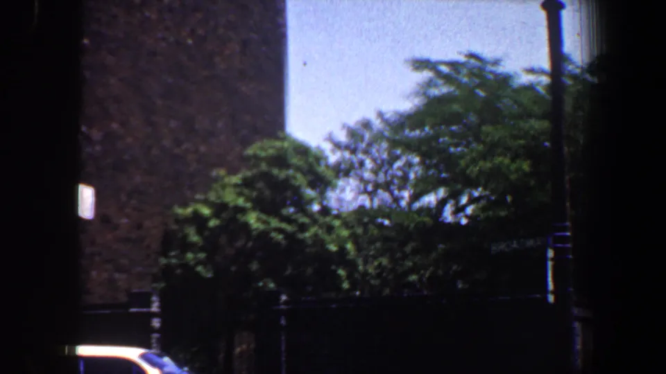 1964 Saint Louis 8mm frame showing a brick building edge, dense green trees, metal railing, and partial dark window area.
